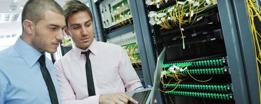 Two men looking at a laptop in front of some servers.